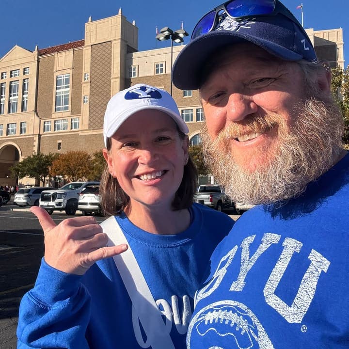 Adam and Allison Benefield in matching blue BYU shirts and caps, smiling in the sun outside a stadium.