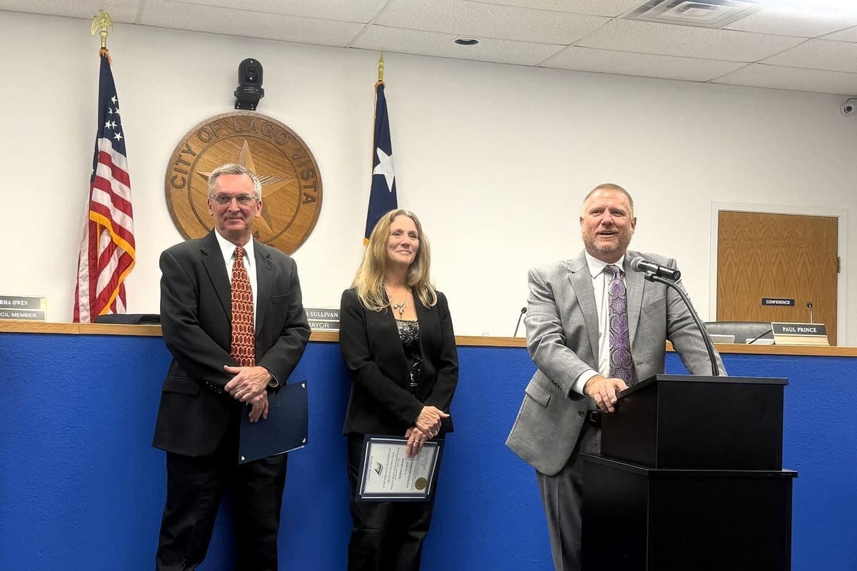 Adam Benefield at the podium in the Lago Vista council chamber, with two other people standing beside the dais and the United States and Texas flags behind them.