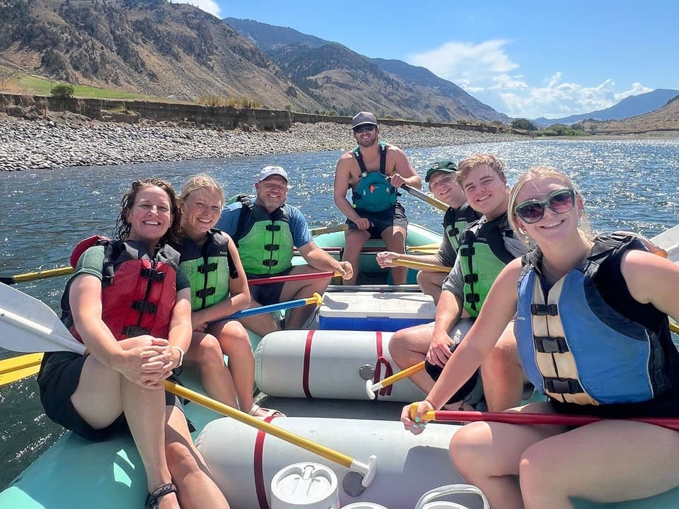Seven members of the Benefield family in life vests on a raft on a river, smiling, with mountains behind them.