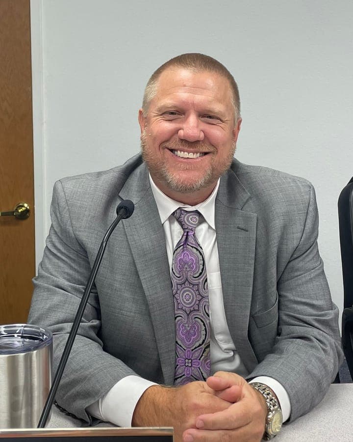 Adam C. Benefield smiling at the council dais in a gray suit and purple patterned tie, hands folded in front of his nameplate.