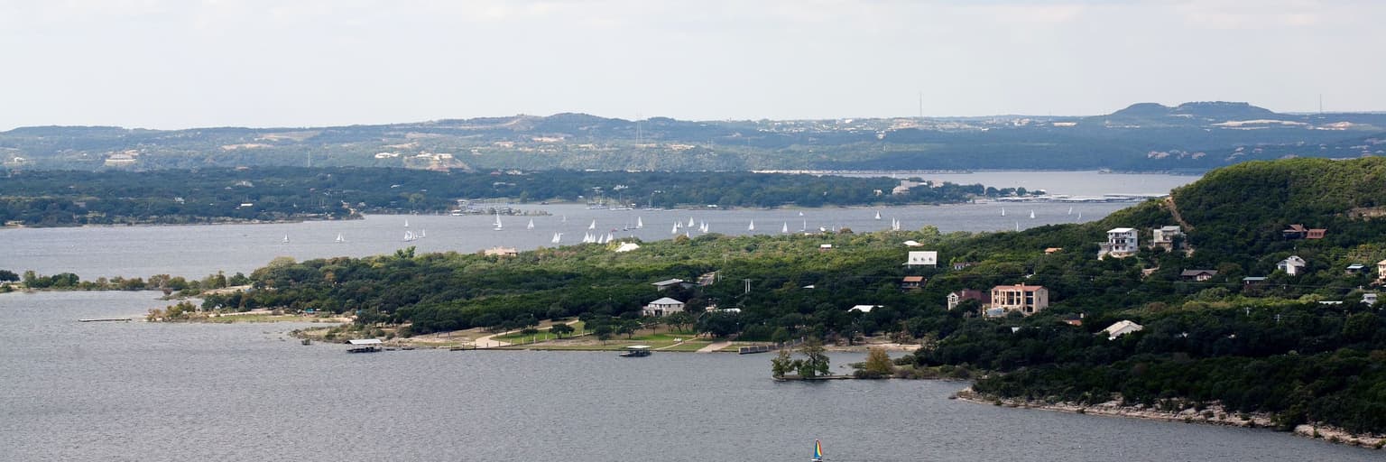 Lake Travis on a bright day: sailboats on the water, a wooded shoreline, and hill-country homes on the far bank.
